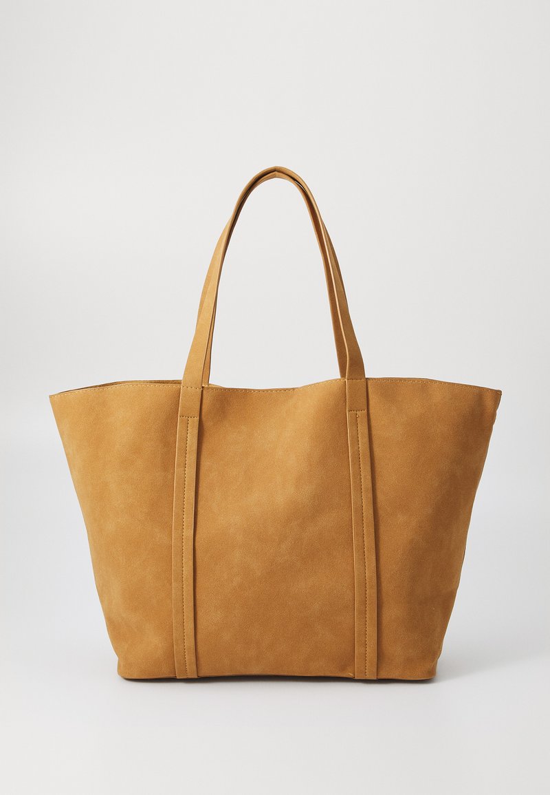 Grand sac fourre-tout beige fabriqué en matériau doux, avec deux anses plates et des détails cousus le long des côtés. Design à ouverture supérieure.