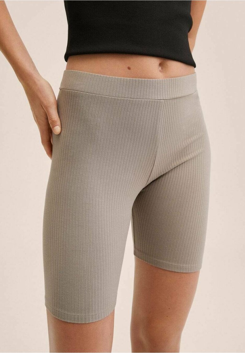 Gros plan d'une personne portant un short cycliste beige, moulant et côtelé, et un top court noir, une main reposant sur sa hanche.