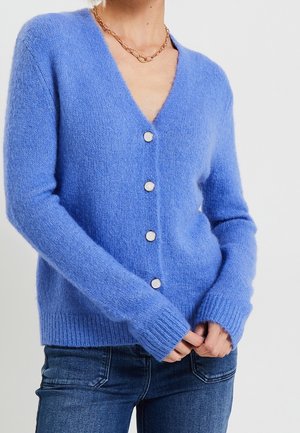 Femme portant un cardigan bleu doux et duveteux boutonné avec un col en V et un collier chaîne doré, associé à un jean bleu foncé.