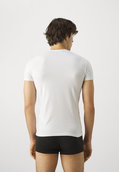 Camiseta blanca de manga corta hecha de una tela ligera y suave, diseño ajustado, escote minimalista, llevada con pantalones cortos negros tipo bóxer, vista desde atrás.