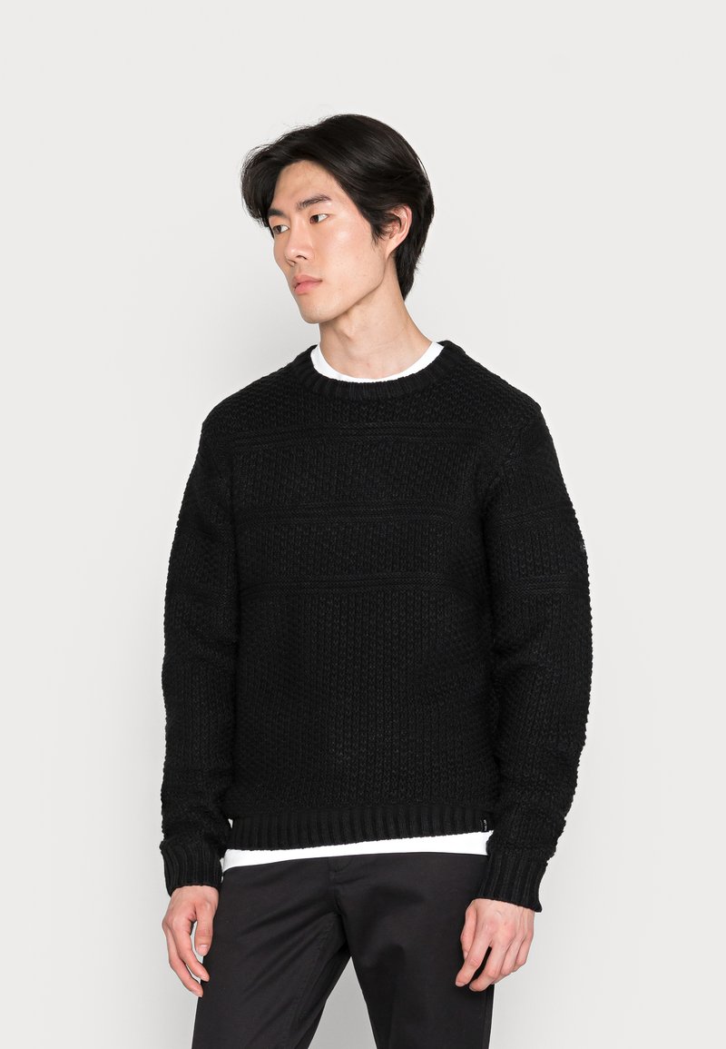 Maglione nero a maglia con un design testurizzato, scollo tondo e polsini e orlo a costine. Indossato sopra una camicia bianca.