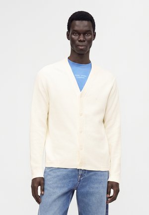 Homme portant un cardigan blanc boutonné par-dessus une chemise bleue et un jean bleu clair sur un fond gris uni.