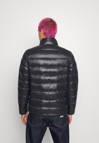 Persona con pelo corto de color rosa brillante que lleva una chaqueta puffer negra brillante y jeans oscuros, de espaldas contra un fondo claro y liso.