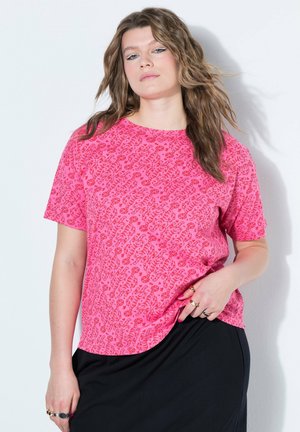 Kobieta o falowanych włosach, ubrana w różową, wzorzystą koszulę z krótkim rękawem i czarną spódnicę, pozująca na białym tle.