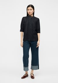 Γυναίκα με μαύρο μπλουζάκι με δαντέλα στο τελείωμα και τρία τέταρτα μανίκια, τζιν παντελόνι με φαρδιά μπατζάκια γυρισμένα προς τα πάνω και καφέ παπούτσια Mary Jane, στέκεται σε λευκό φόντο.