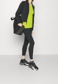 Chaqueta deportiva negra, camiseta verde neón, leggings negros y zapatillas negras con acentos blancos. El modelo sostiene un bolso negro. Fondo simple.