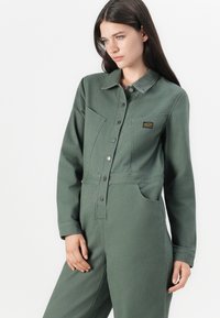 Jumpsuit en denim vert avec des manches longues, col classique, fermeture à boutons sur le devant et deux poches poitrine. Présente un petit patch logo sur la poitrine.