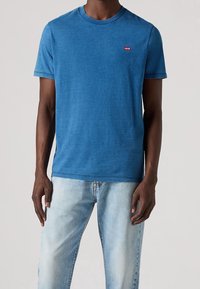 Modré tričko s kulatým výstřihem vyrobené z bavlny s hladkou texturou; má malé červené logo Levi's na levé straně hrudi a krátké rukávy.