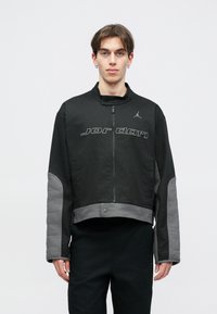 Casaco cropped preto com acabamentos cinzas, apresenta um colarinho alto, fecho com zíper, mangas acolchoadas e o logótipo "Jordan" no peito.