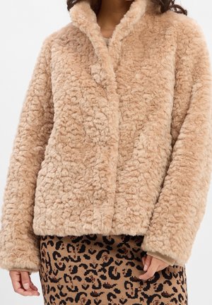 Femme portant une veste en fausse fourrure duveteuse marron clair sur une jupe beige et noire à imprimé léopard, les mains détendues le long du corps.