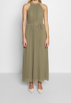 Kvinde iført ærmeløs, olivengrøn, plisseret maxikjole med bindebånd i taljen og beige spidse hæle mod ensfarvet baggrund.