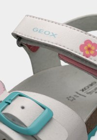 Λευκό παιδικό σανδάλι με μπλε αγκράφα, ροζ διακόσμηση με λουλούδι και λογότυπο "GEOX" στο λουράκι του αστραγάλου.