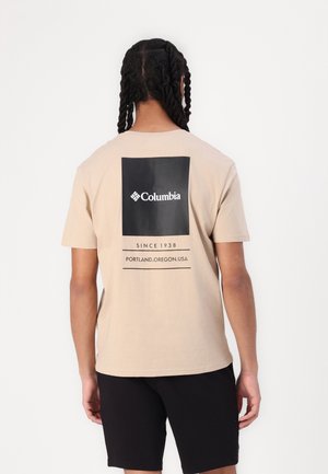 Personne aux cheveux tressés portant un t-shirt Columbia beige avec un logo noir et le texte "Depuis 1938 Portland, Oregon, USA", associé à un short noir.