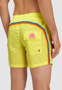 Pantaloni da bagno giallo brillante con tasca sul retro, caratterizzati da strisce laterali multicolori blu, arancione e rosso, realizzati in tessuto leggero.