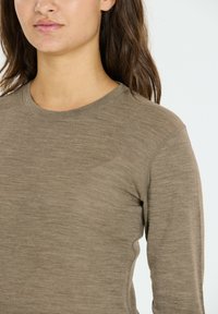 Pitkähihaisen paidan muted ruskeasta väristä, joka on valmistettu kevyestä, teksturoidusta kankaasta, pyöreällä pääntiellä ja vartalonmyötäisellä leikkauksella. Saumadetailit hartioilla.