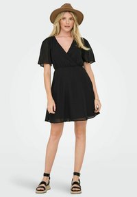 Robe noire portefeuille avec des manches courtes en volant, une taille cintrée et une jupe évasée. Tissu lisse et décolleté en V. Associée à un chapeau marron et des sandales noires.