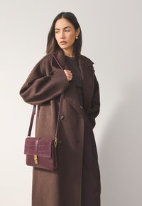 Brown oversized coat with large lapels, paired with a textured burgundy crossbody bag featuring a gold clasp and embossed pattern.