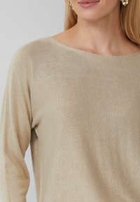 Beige gestrickter Pullover mit weitem Halsausschnitt, der dezente metallische Fäden für einen schimmernden Effekt aufweist. Glatte Textur, lange Ärmel.