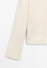 Maglione lavorato a maglia in bianco sporco con una texture a coste. Presenta una tasca sul lato sinistro e polsini a coste per una vestibilità strutturata.