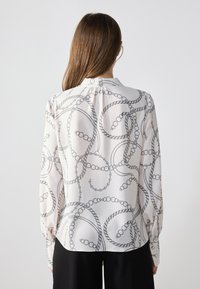 Blusa a maniche lunghe in tessuto leggero e setoso con un motivo a catena nera su fondo crema; include un colletto classico e chiusura a bottoni.