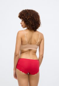 Μπεζ αόρατο σουτιέν με ρυθμιζόμενους ιμάντες, συνδυασμένο με κόκκινες εσώρουχες με δαντέλα. Οι υφές είναι λείες και μαλακές.