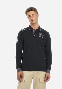 Polo a maniche lunghe di colore blu navy con colletto e polsini grigi chiari, con loghi e testi ricamati in bianco sul lato sinistro del petto e sulla spalla.