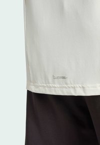 Bílý sportovní top s jemnou texturou, na kterém se nachází štítek "CLIMACOOL" blízko lemu. Spodní část je tmavá, čímž vytváří dvoubarevný efekt.
