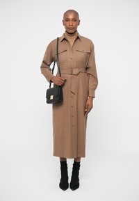 Robe marron à manches longues, à boutons, avec une ceinture, deux poches poitrine, associée à un petit sac noir et des bottes noires à bout pointu.