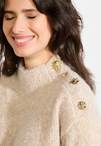 Bežs adīts džemperis ar augstu apkakli, ar trim dekoratīvām zelta pogām plecu daļā un mīkstu, teksturētu audumu.