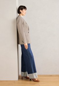 Double-breasted houndstooth blazer in beige, wide-leg denim pants with frayed hem, and brown high-heeled shoes. Standing against a wall.