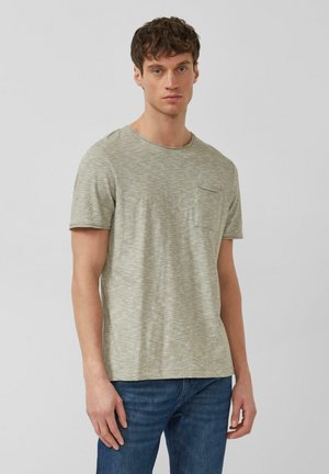 Junger Mann steht, trägt ein hellgraues T-Shirt mit kurzen Ärmeln und Brusttasche sowie eine blaue Jeans vor einfarbig grauem Hintergrund.