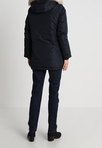 Zwarte pufferjas met een bontgevoerd capuchon, gestikte textuur en een middellange uitstraling. Draagt een combinatie met donkere jeans en enkellaarsjes.