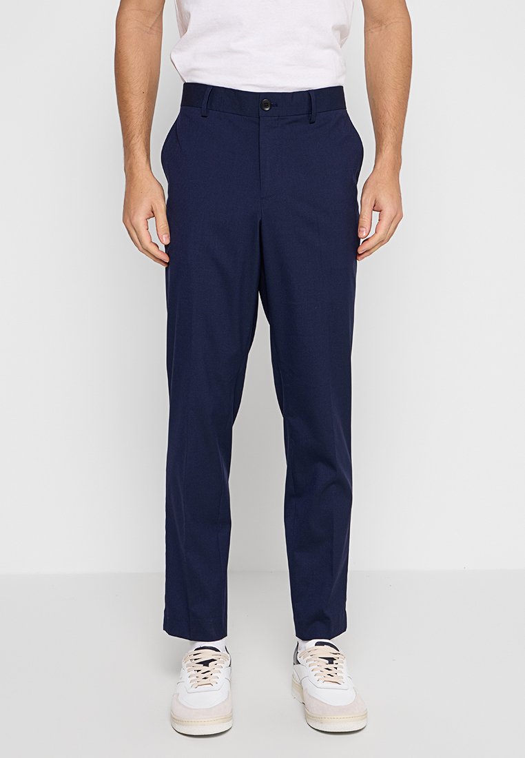 Marineblaue maßgeschneiderte Hosen aus glattem Stoff mit klassischem Schnitt, Fronttaschen und Knopfverschluss, getragen mit weißen Sneakers.