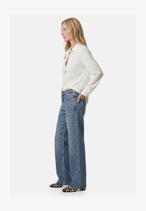 Nainen seisoo sivuttain, yllään valkoinen napitettava paita, leveälahkeiset siniset farkut ja leopardikuviolliset teräväkärkiset ballerinakengät.