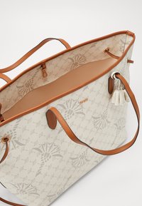 Canvas toteväska med en beige och ljusbrun mönstrad utsida, med bruna läderhandtag, en tanfärgad insida och dekorativa tofsar.
