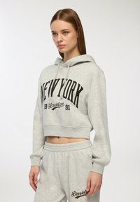 Szara krótka bluza z kapturem z czarnym nadrukiem "NEW YORK", z regulowanym kapturem. W komplecie z pasującymi spodniami dresowymi w tym samym odcieniu szarości, z detalem logo.