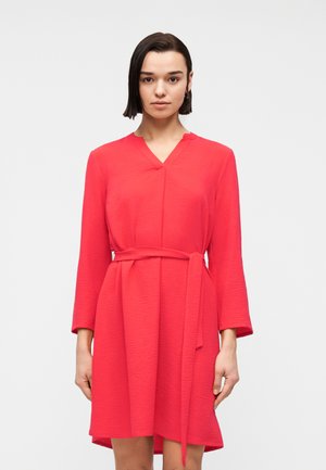 Kvinne iført en knallrød, langermet kjole med V-hals og matchende belte, stående mot en ensfarget lys bakgrunn.