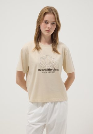 Jeune femme portant un t-shirt beige avec un motif coquillage et le texte « Beach Rhythm Feel the Beach Beat », associé à un pantalon blanc.