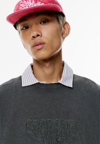 Grauer Sweatshirt mit abgenutztem Kragen, über ein weiß gestreiftes Hemd getragen. Die Person trägt eine pinke Mütze mit besticktem Text.