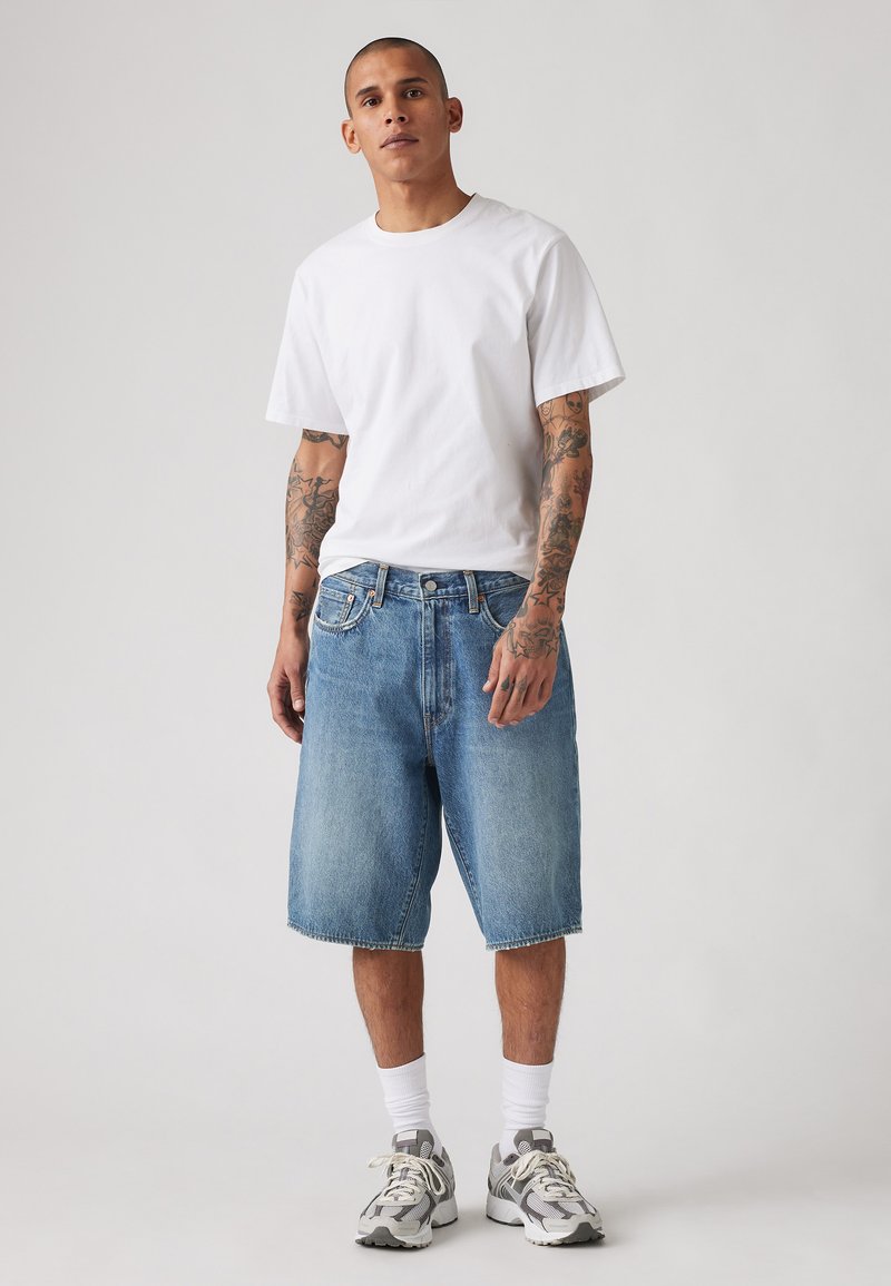 Light blue denim shorts with a relaxed fit, paired with a plain white cotton t-shirt and grey sneakers. Tattoos are visible on arms.