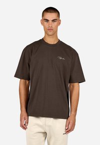 Camiseta holgada de algodón marrón con cuello redondo y mangas cortas, que presenta un pequeño logo blanco bordado en el pecho izquierdo.