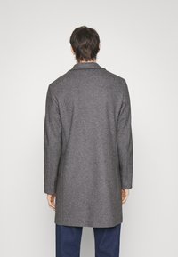 Uomo di spalle che indossa un lungo cappotto di lana grigia e pantaloni blu su uno sfondo semplice chiaro.