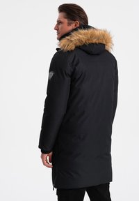 Parka nera imbottita con rifinitura in pelliccia sintetica sul cappuccio, tasche con zip posteriori e patch con logo sul braccio sinistro. Texture morbida e design di lunghezza media.