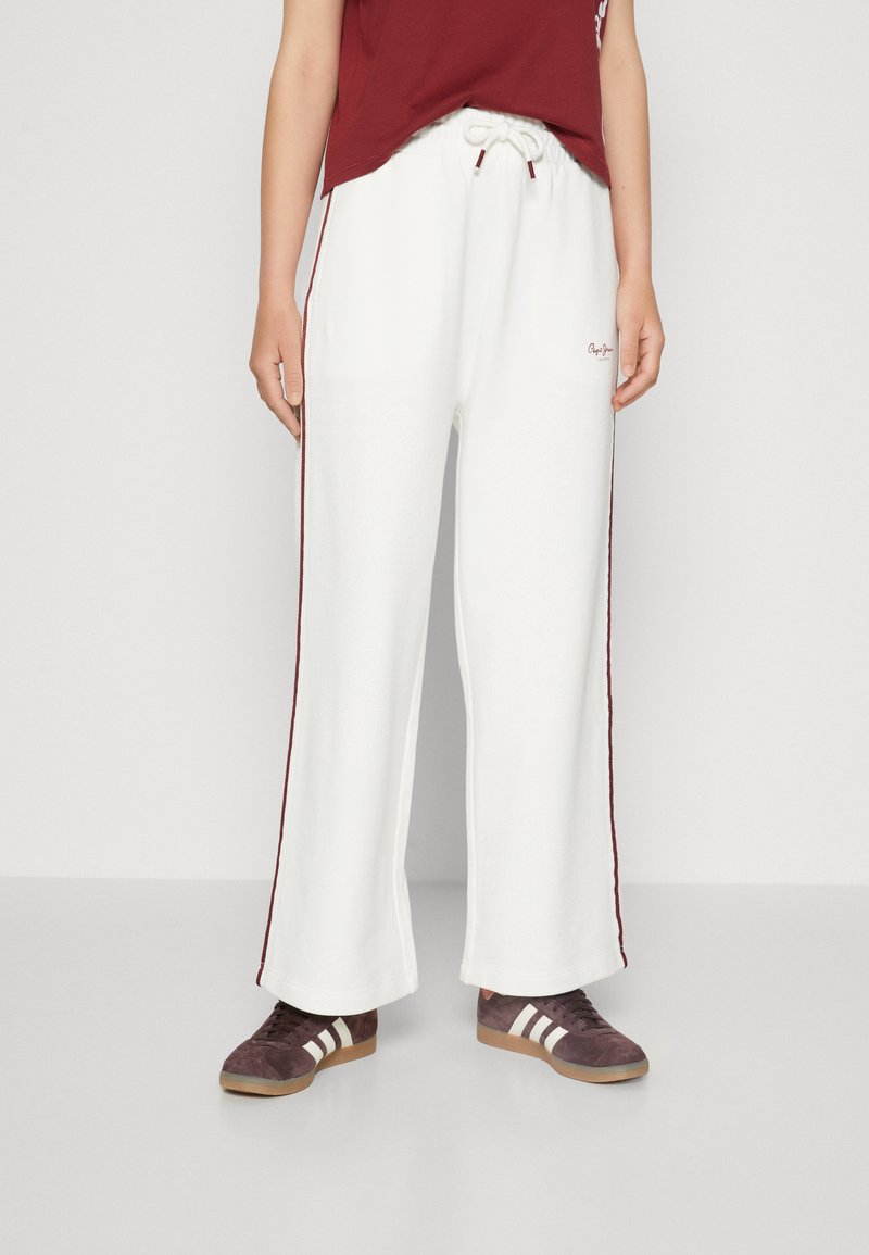 Pantaloni larghi bianchi con vita regolabile, dotati di strisce laterali rosso scuro e logo ricamato. Indossati con scarpe sportive marroni.