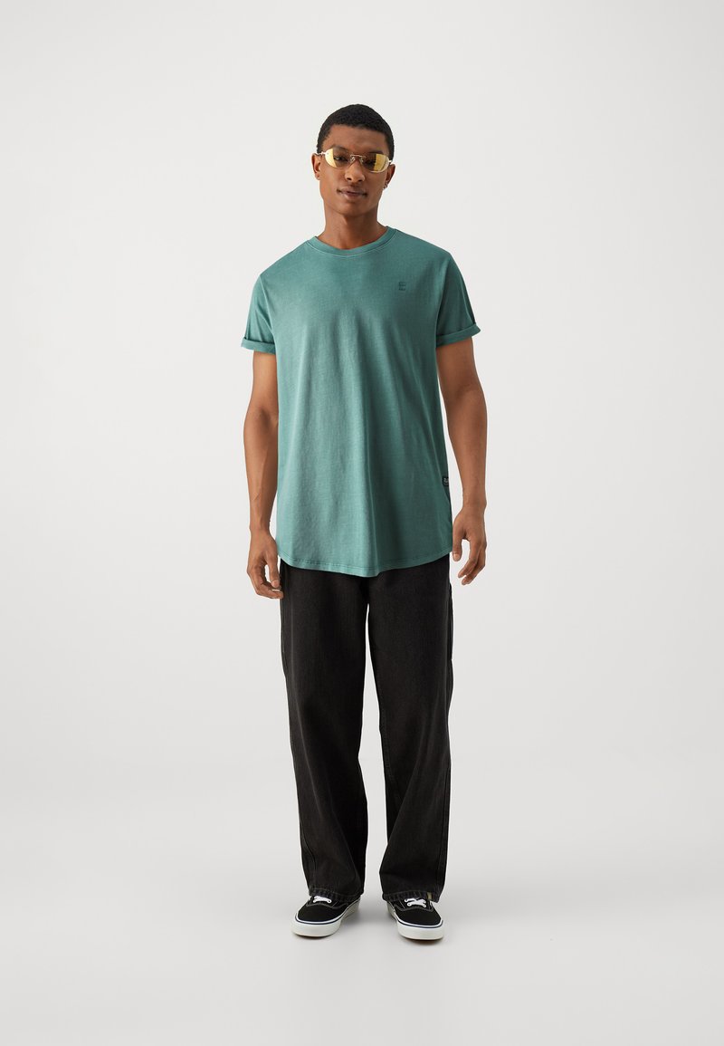 Türkisfarbene Baumwoll-T-Shirt mit umgeschlagenen Ärmeln, getragen mit schwarzen, weit geschnittenen Hosen und schwarzen Sneakers. Das Model trägt eine gelbe Sonnenbrille und steht vor einem schlichten Hintergrund.