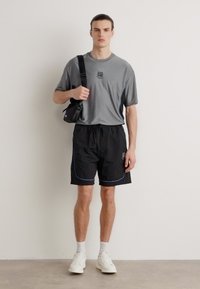 Camiseta deportiva corta de color gris con un logo, pantalones cortos negros con detalles azules y zapatillas blancas. Modelo sosteniendo una pequeña bolsa negra.