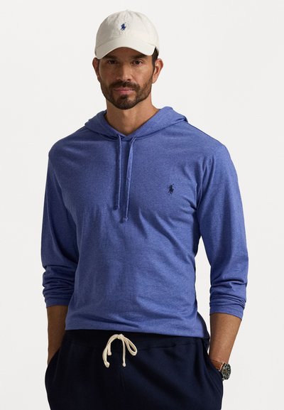 Hanorac albastru cu mâneci lungi și șnur, cu un logo mic pe piept. Poartă cu șorturi bleumarin și o pălărie de culoare deschisă.