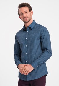 Camicia navy con bottoni, a maniche lunghe e collo a punta, decorata con piccoli pois bianchi e rossi. Il tessuto appare liscio e strutturato.