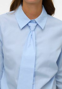 Jasnoniebieska koszula na guziki z wyraźnym kołnierzem i krawatem na całej długości. Tkanina wydaje się gładka i lekko błyszcząca. Zbliżenie na linię szyi.