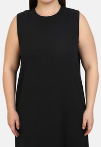 Mustaa hihaton mekko, jossa on pyöreä pääntie ja sileä, teksturoitu materiaali. Mekko on löysä ja ulottuu juuri vyön ylle.